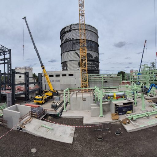 Weitläufige Industriebaustelle mit Stahlkonstruktionen, Kränen, Hubarbeitsbühnen und einem großen zylindrischen Industriegebäude im Hintergrund.