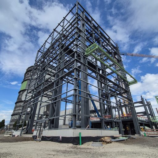 Stahlkonstruktion eines mehrstöckigen Industriegebäudes im Bau vor blauem Himmel, mit sichtbaren Tragwerken und Montageelementen.