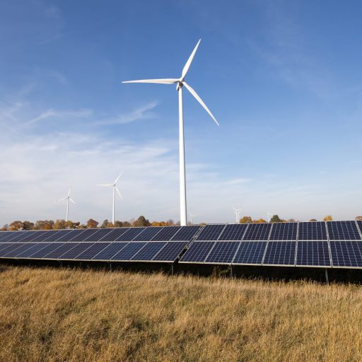 Luftaufnahme einer weitläufigen Landschaft mit Photovoltaikanlagen und Windkraftanlagen auf grünen Feldern unter einem Himmel mit Wolken – Symbolbild für erneuerbare Energien und Nachhaltigkeit.