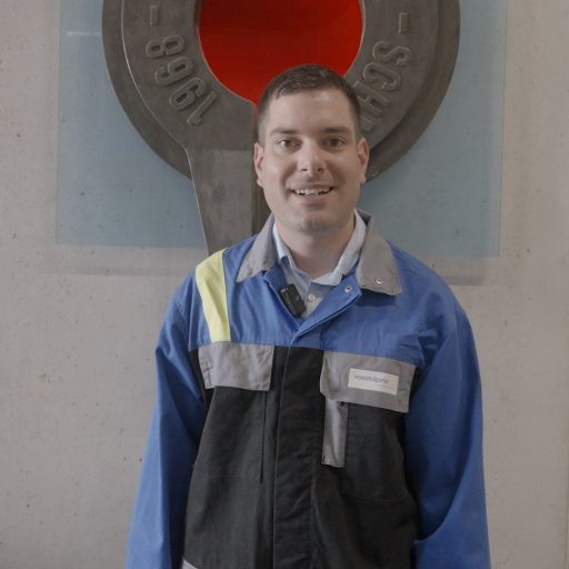 Christian H. aus dem voestalpine Forschungsteam steht in Arbeitskleidung vor einer Werkshallenwand – bereit für den Einsatz in der Werkstoffanalyse.