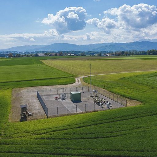Aufnahme der Forschungsanlage Rubensdorf, eingezäunt und inmitten von landwirtschaftlich bewirtschafteten Äckern. Am Horizont sind Häuser und Berge zu sehen.