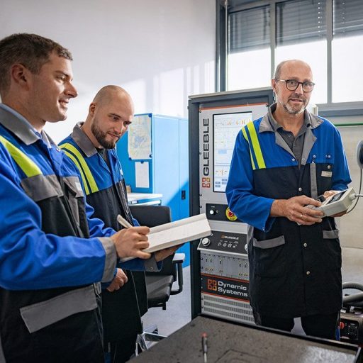Drei voestalpine Mitarbeiter in Arbeitskleidung besprechen Messergebnisse vor einem Gleeble-Prüfgerät im Laborumfeld. Einer hält ein Notizbuch, ein anderer ein Steuergerät.