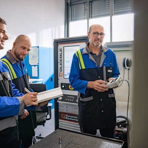 Drei voestalpine Mitarbeiter in Arbeitskleidung besprechen Messergebnisse vor einem Gleeble-Prüfgerät im Laborumfeld. Einer hält ein Notizbuch, ein anderer ein Steuergerät.