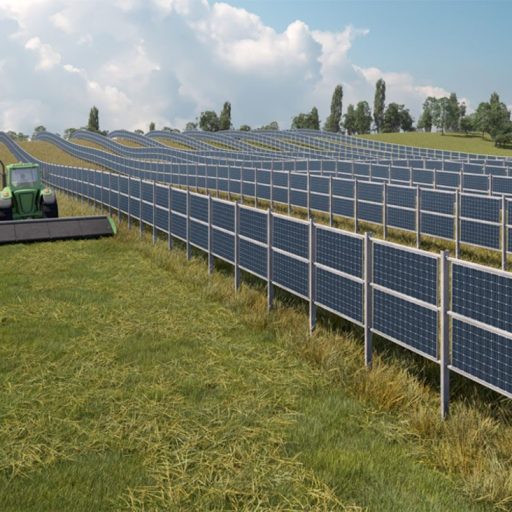 Ein grüner Traktor mäht Gras zwischen erhöhten PV-Modulen auf einer großen Solaranlage in einer Wiese, umgeben von Bäumen und blauem Himmel.