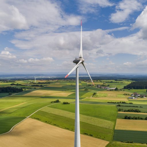 A large wind turbine towers over a green, patchwork-like agricultural landscape with fields, small villages and other wind turbines under a partly cloudy sky.