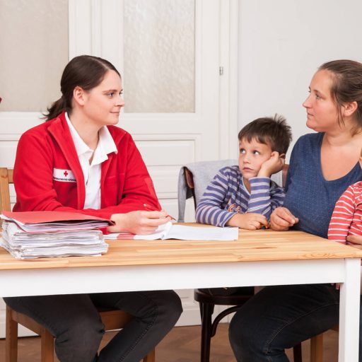 Eine Mutter mit Ihrem Sohn und ihrer kleinen Tochter am Schoß spricht sitzt am Schreibtisch einer Rotes Kreuz Mitarbeiterin