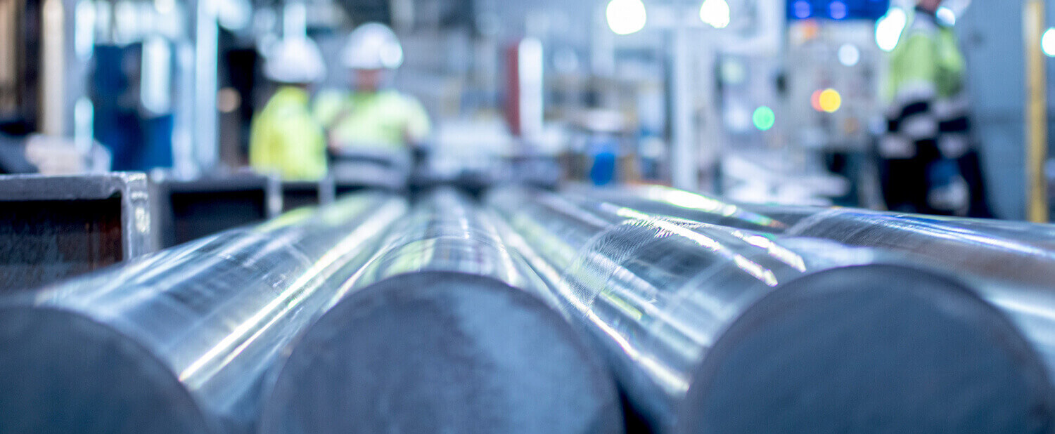 Close-up view of large tool steel rods in a factory setting, with two workers in high-visibility clothing and hard hats blurred in the background. Bright industrial lighting highlights the shiny metal surfaces ideal for tooling applications.