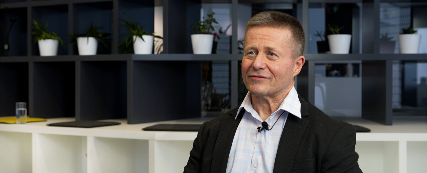 Matko Bozanic in a suit and open-collared shirt sits in a modern office with shelves, potted plants, and a clock in the background. He is looking slightly to the side and appears to be in conversation or being interviewed.