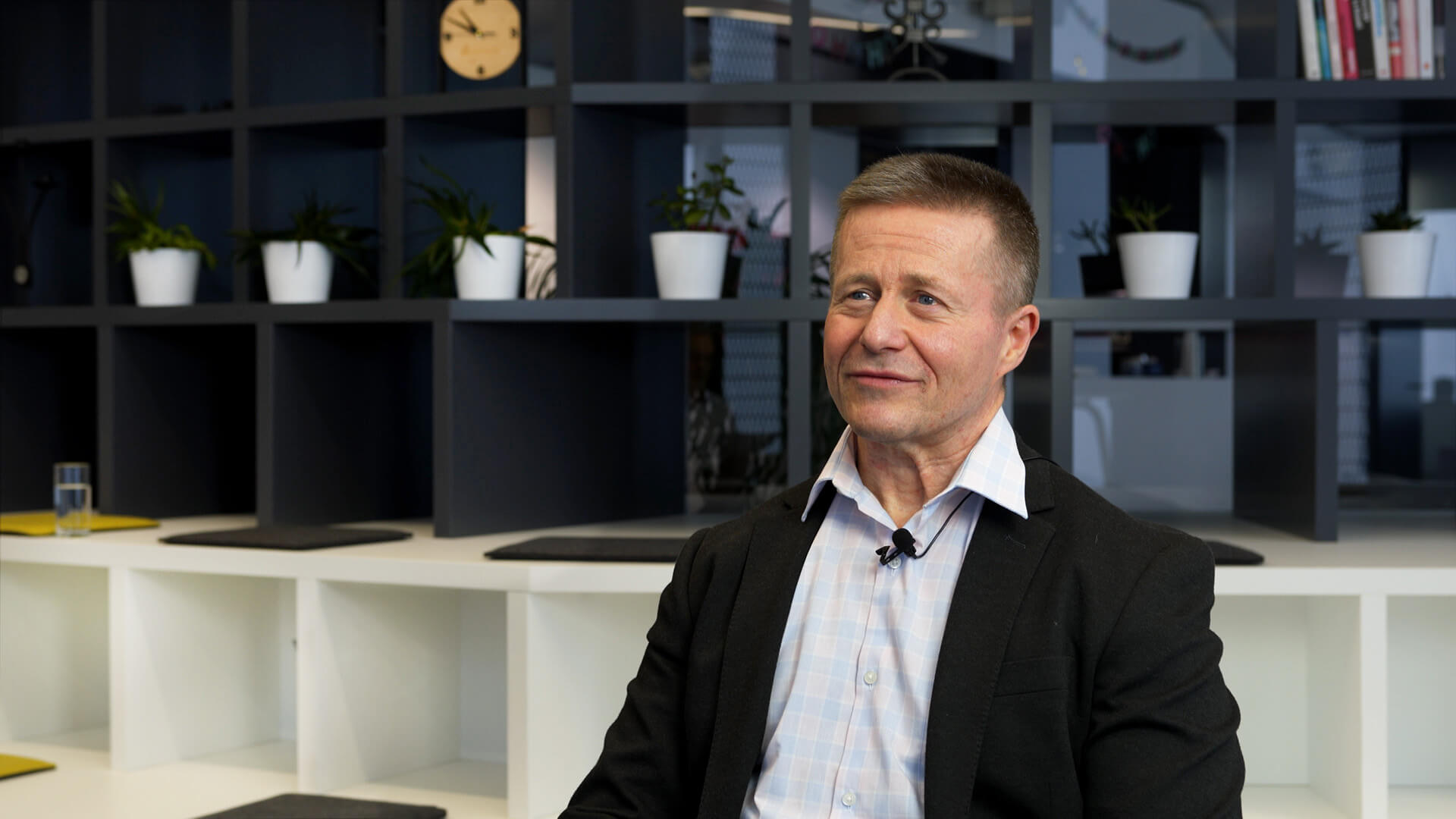 Matko Bozanic in a suit and open-collared shirt sits in a modern office with shelves, potted plants, and a clock in the background. He is looking slightly to the side and appears to be in conversation or being interviewed.