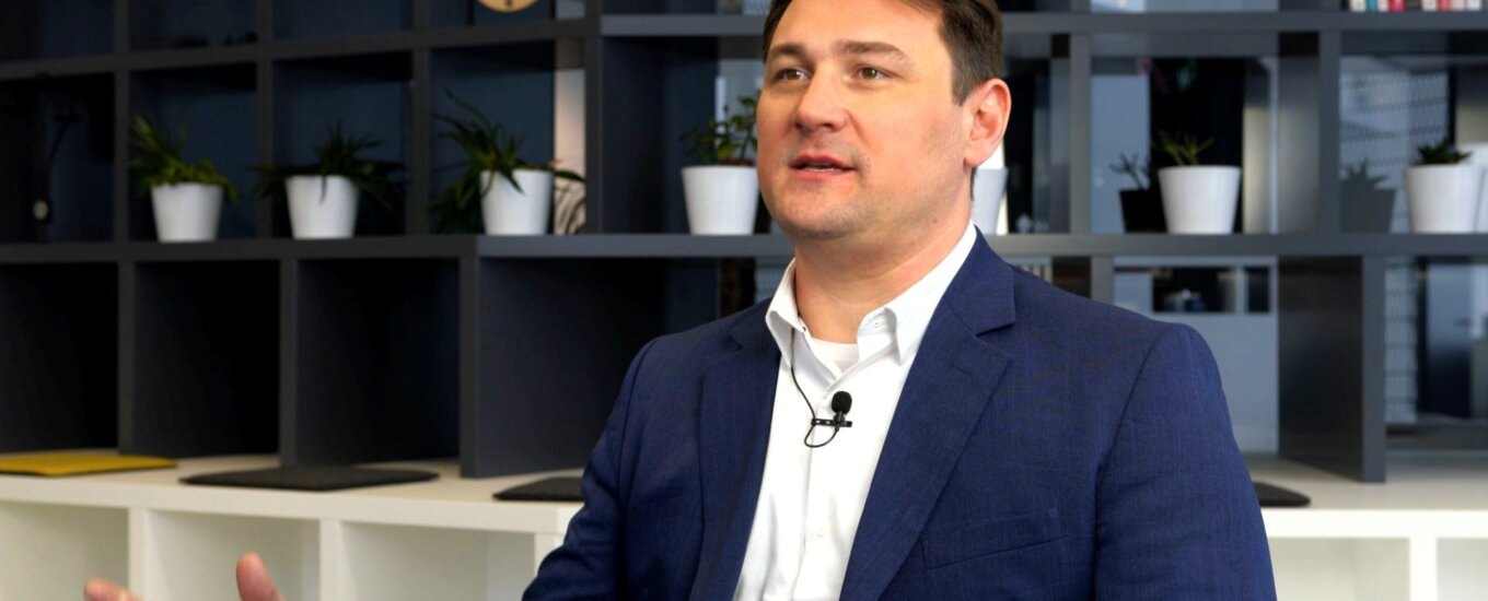 A man in a dark blue suit and white shirt gestures while speaking, sitting in a modern office with shelves, potted plants, books, and a clock in the background.