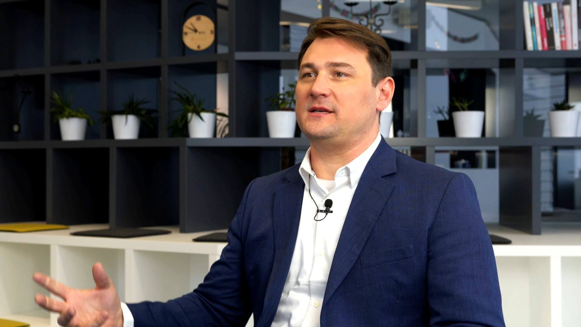 A man in a dark blue suit and white shirt gestures while speaking, sitting in a modern office with shelves, potted plants, books, and a clock in the background.