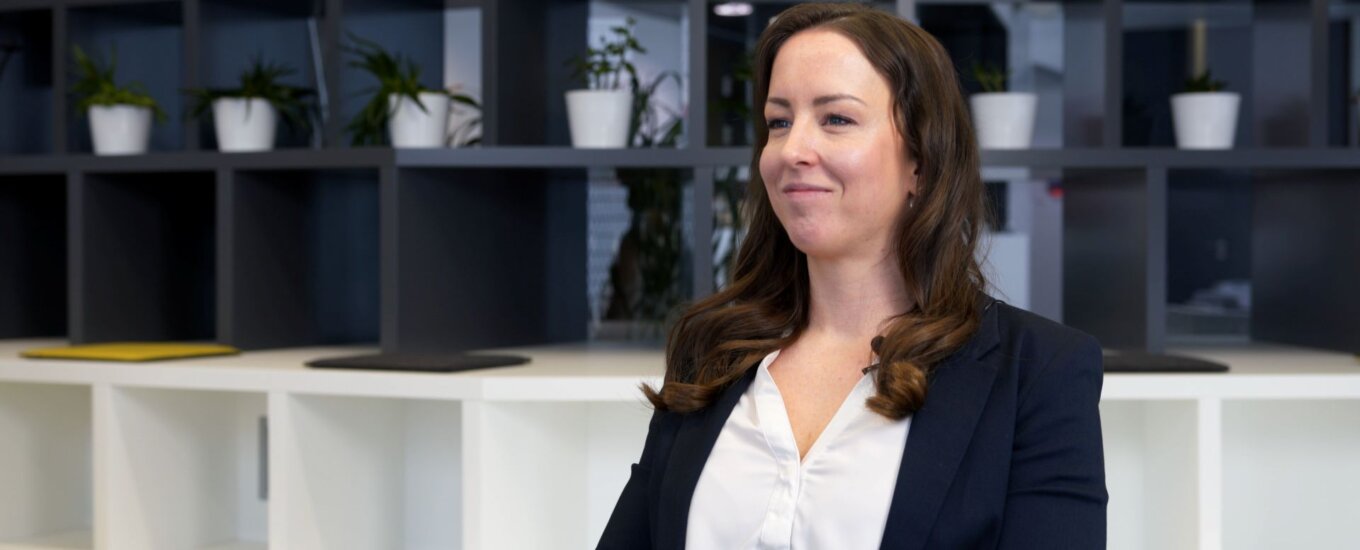 Carina Weninger: A woman with long brown hair, wearing a dark blazer and white blouse, sits and smiles in a modern office with shelves, books, plants, and a clock in the background.