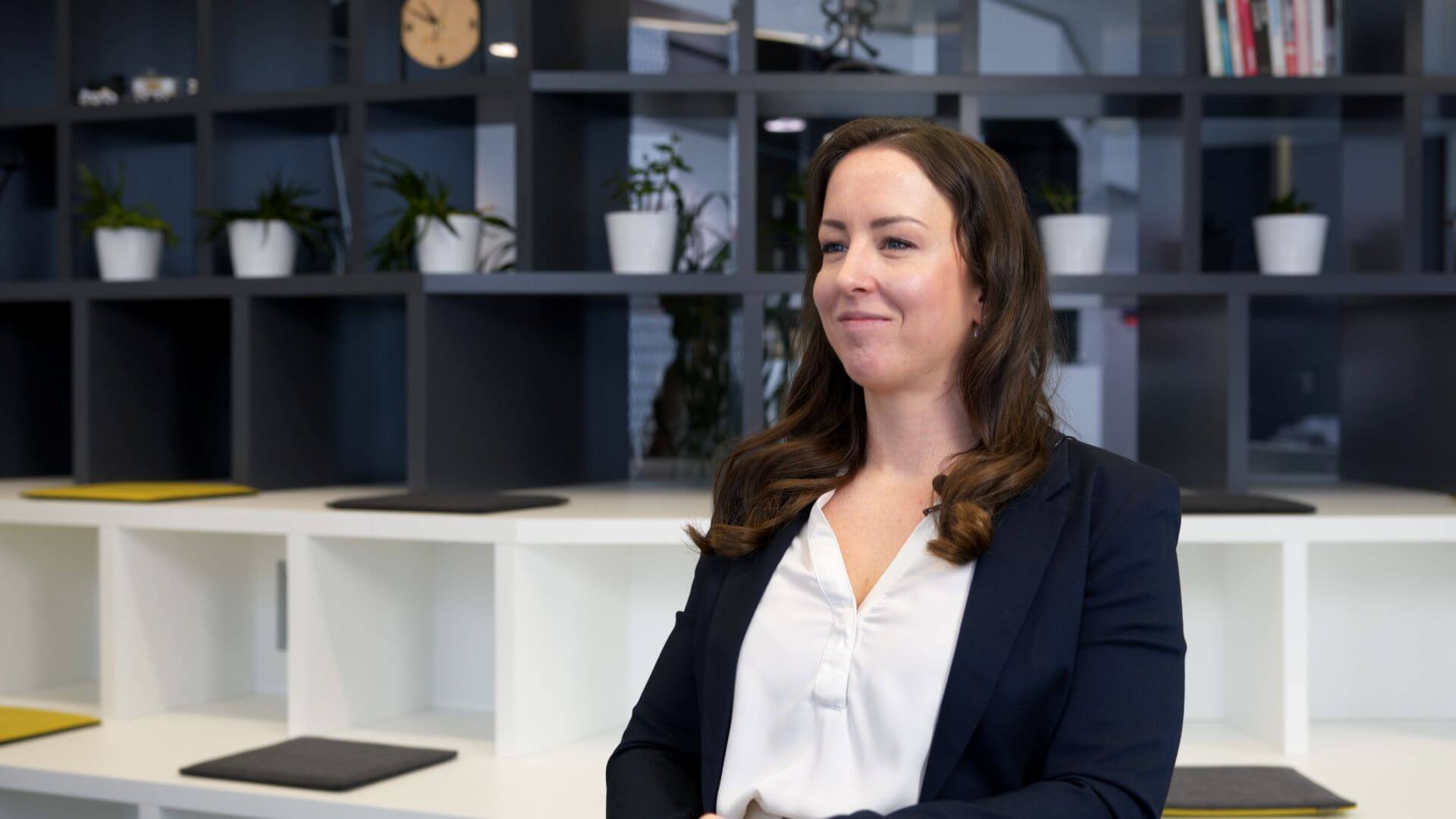 Carina Weninger: A woman with long brown hair, wearing a dark blazer and white blouse, sits and smiles in a modern office with shelves, books, plants, and a clock in the background.