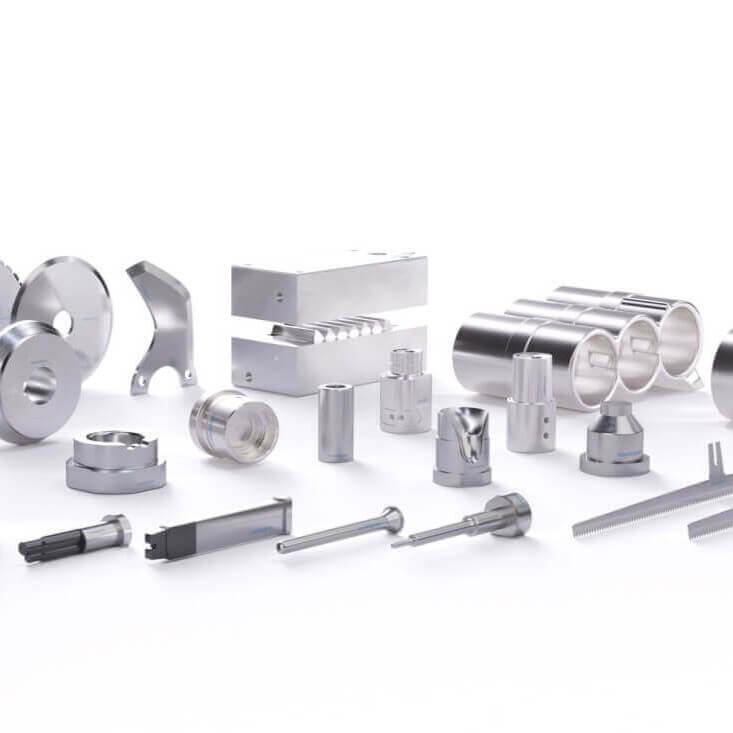 Eine Sammlung verschiedener glänzender maschinell bearbeiteter Metallteile und Komponenten, darunter Zylinder, Zahnräder, Blöcke und Stangen, die auf einer weißen Fläche mit neutralem Hintergrund angeordnet sind.