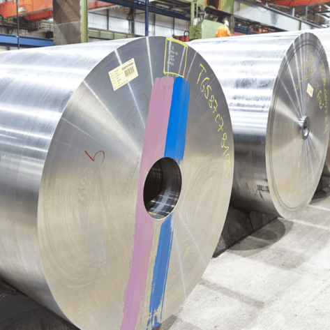 Große, glänzende Rollen mit Etiketten und farbigen Markierungen sind in einer Fabrikhalle gestapelt und spiegeln das Licht der Oberlichter in einer industriellen Umgebung wider.