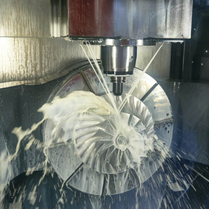 Eine CNC-Fräsmaschine bearbeitet ein Turbinenteil aus Metall, wobei eine Kühlflüssigkeit über das sich drehende Bauteil gesprüht wird, um den Zerspanungsprozess zu kühlen und zu schmieren.