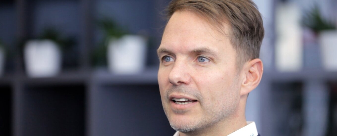 Dominik Baumann, with light brown hair and blue eyes, wearing a dark sweater and a white shirt,, sits indoors with shelves and potted plants blurred in the background.
