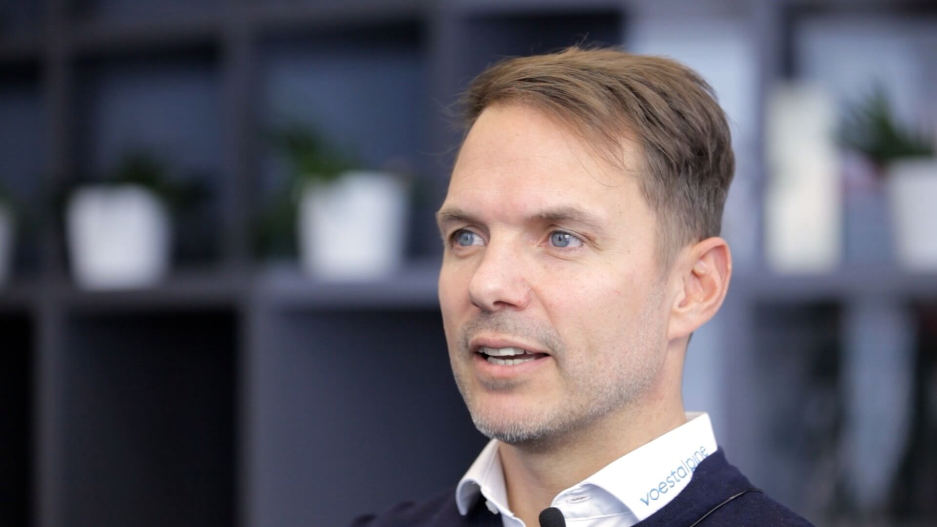 Dominik Baumann, with light brown hair and blue eyes, wearing a dark sweater and a white shirt,, sits indoors with shelves and potted plants blurred in the background.