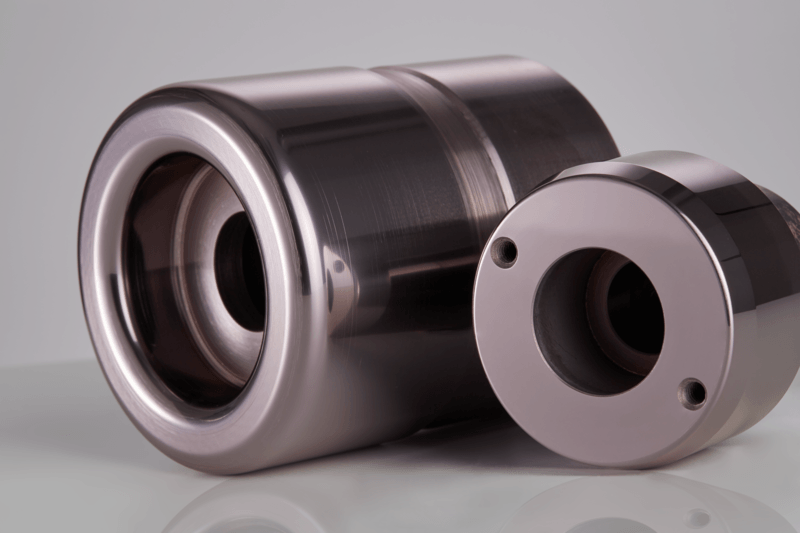 Two shiny metallic cylindrical machine parts with smooth surfaces and hollow centers are positioned on a reflective surface against a neutral background.