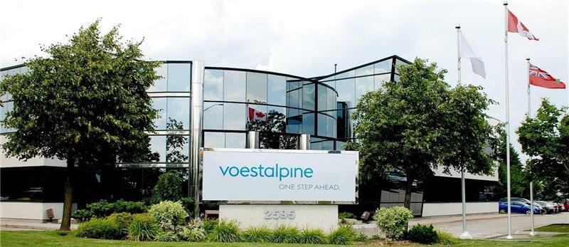 A modern office building with reflective windows, trees, two flagpoles, and a large sign reading voestalpine ONE STEP AHEAD. 2958 in front. The Canadian flag and another flag are visible.