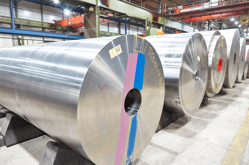 Large rolls of shiny aluminum sheet metal and engineering steels are lined up on supports inside a brightly lit industrial warehouse, with colored markings and labels visible on the ends of the rolls.