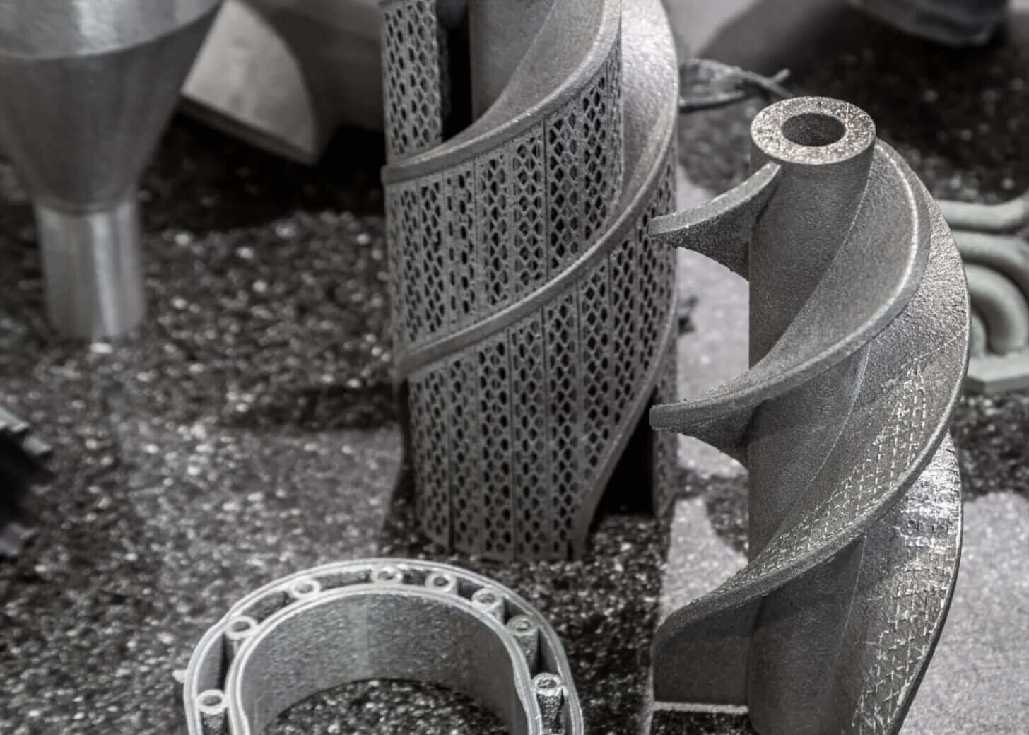 Various metallic 3D-printed industrial components with intricate shapes and hollow sections are arranged on a textured, dark surface, highlighting complex geometric designs and fabrication techniques.