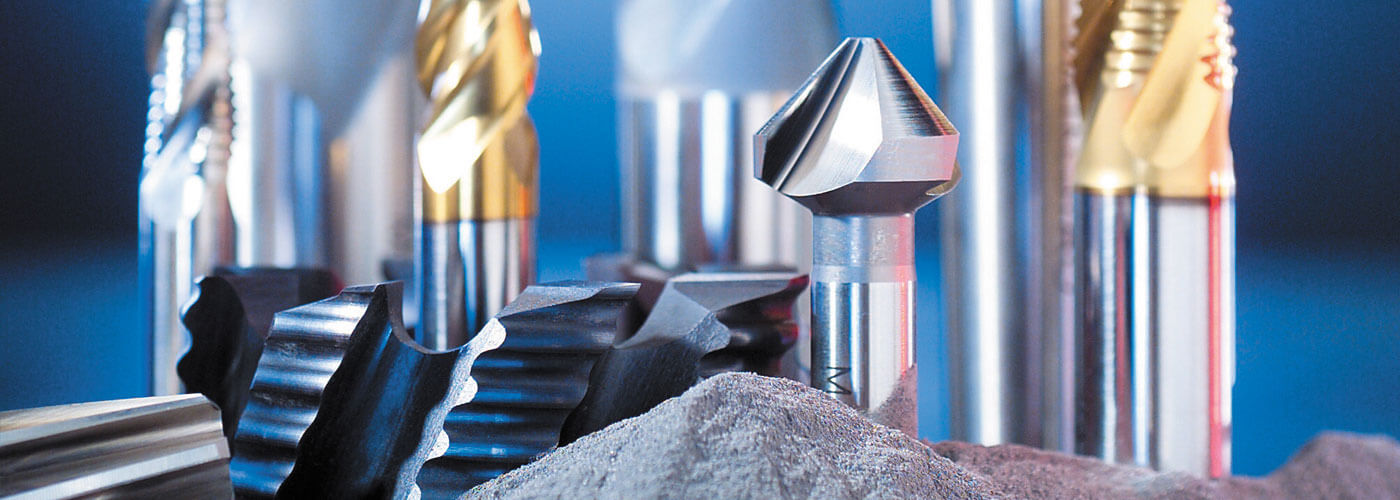 A close-up view of various high speed steel cutting tools and drill bits standing upright in a group, with a mound of metallic shavings or powder in the foreground, set against a blue background.