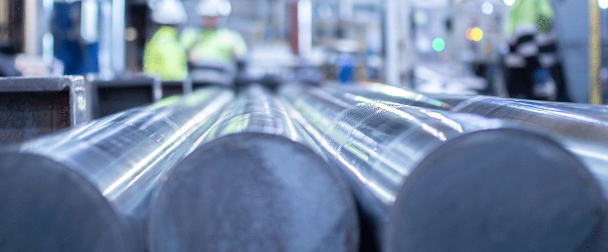 Primer plano de varias barras metálicas de gran tamaño enfocadas, con trabajadores con equipo de seguridad y un entorno de fábrica difuminado al fondo.