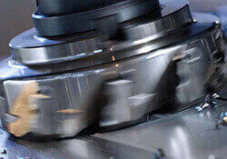 Close-up of a metal gear in motion, cutting through a metallic surface with small metal shavings scattered around, demonstrating a machining or milling process.