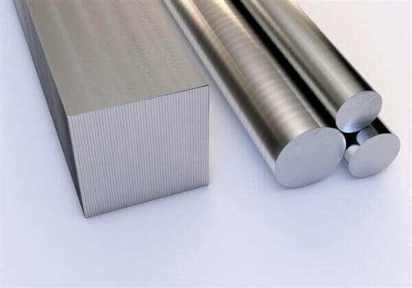 A rectangular metal bar and three cylindrical metal rods are placed next to each other on a smooth, white surface. The materials have a shiny, reflective, metallic finish.
