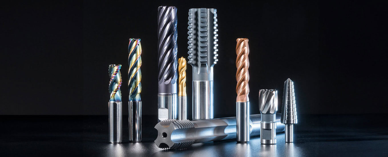 A group of various metal cutting tools, including high speed steel drill bits, end mills, and taps with different shapes, sizes, and coatings, arranged upright on a dark surface against a black background.