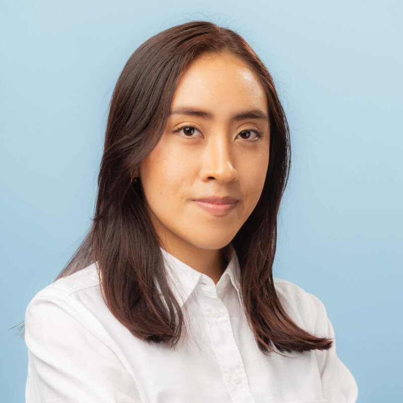 A woman with straight brown hair, wearing a white button-up shirt, poses confidently in front of a plain light blue background, ready for contacto or networking opportunities.