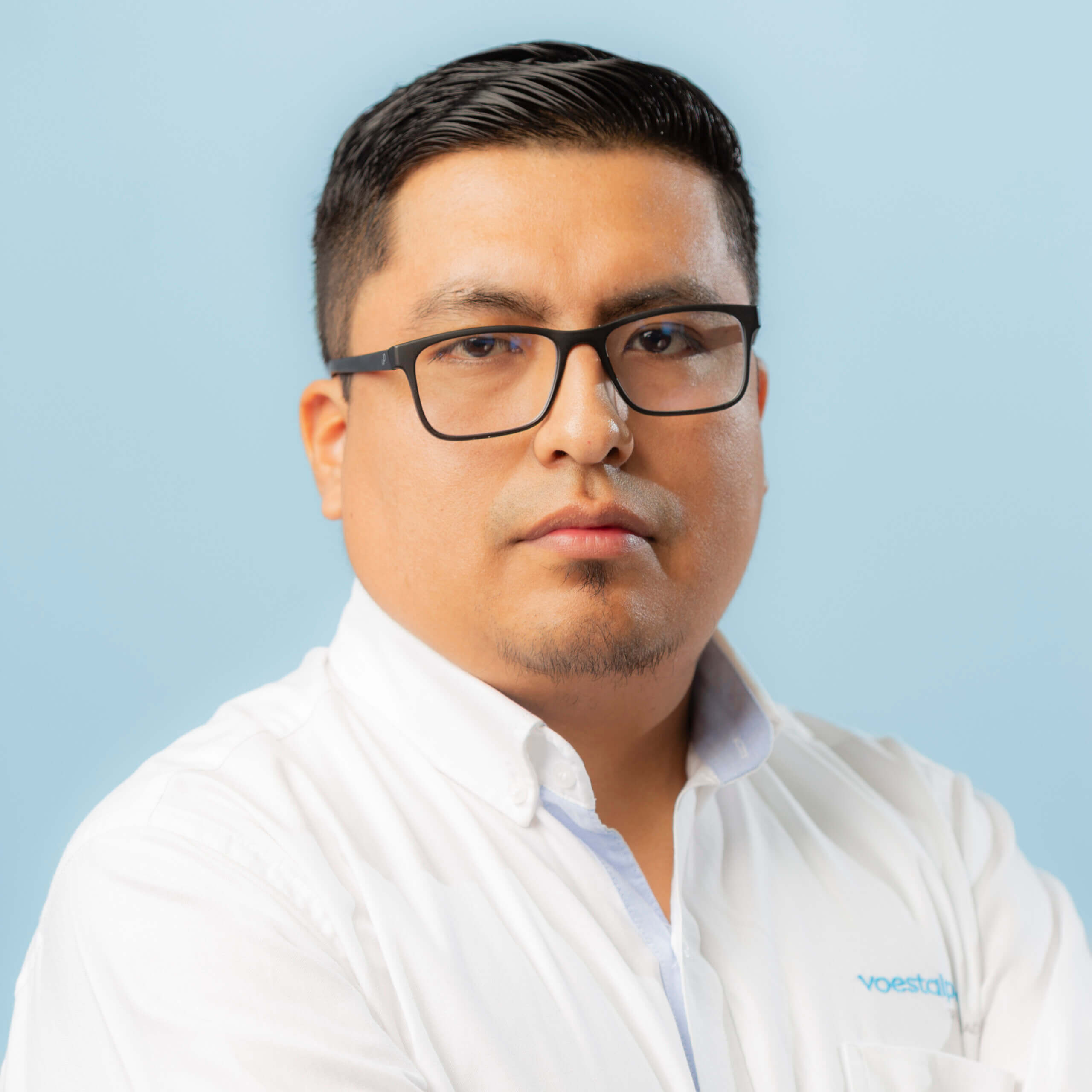 A man with short black hair, glasses, and a trimmed goatee wearing a white collared shirt stands against a light blue background with a neutral expression.