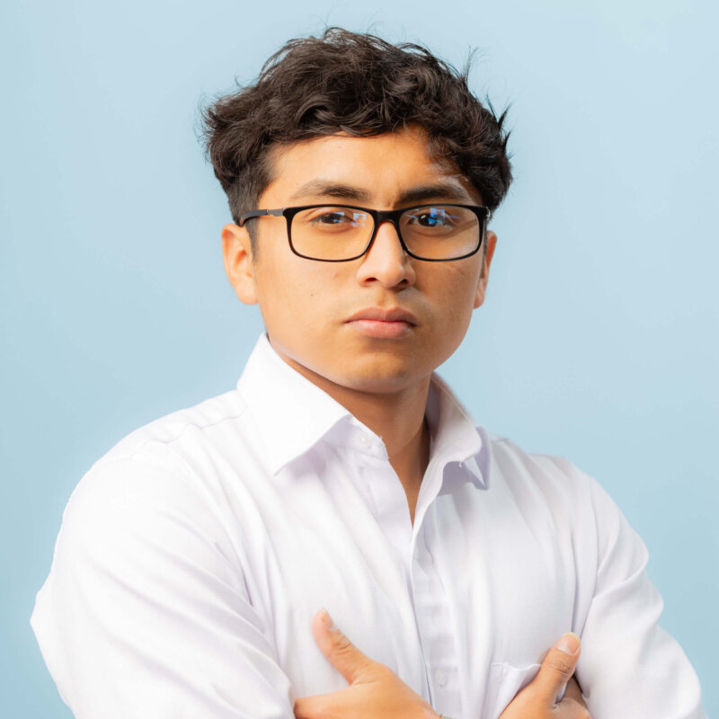 A young man with short dark hair and glasses, wearing a white button-up shirt, stands with arms crossed against a light blue background, looking confidently at the camera.