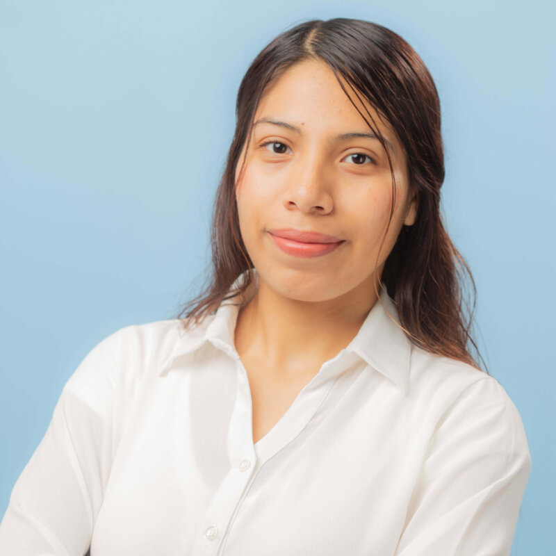 A young woman with long dark hair and a light complexion, wearing a white collared shirt, stands against a light blue background, smiling softly with her arms crossed.