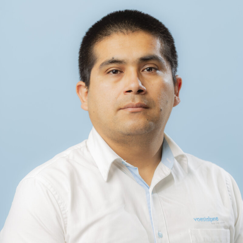 A man with short dark hair wearing a white collared shirt stands against a light blue background. He faces the camera with a neutral expression. The shirt, featuring a small blue logo on the chest, is perfect for making un contacto profesional.