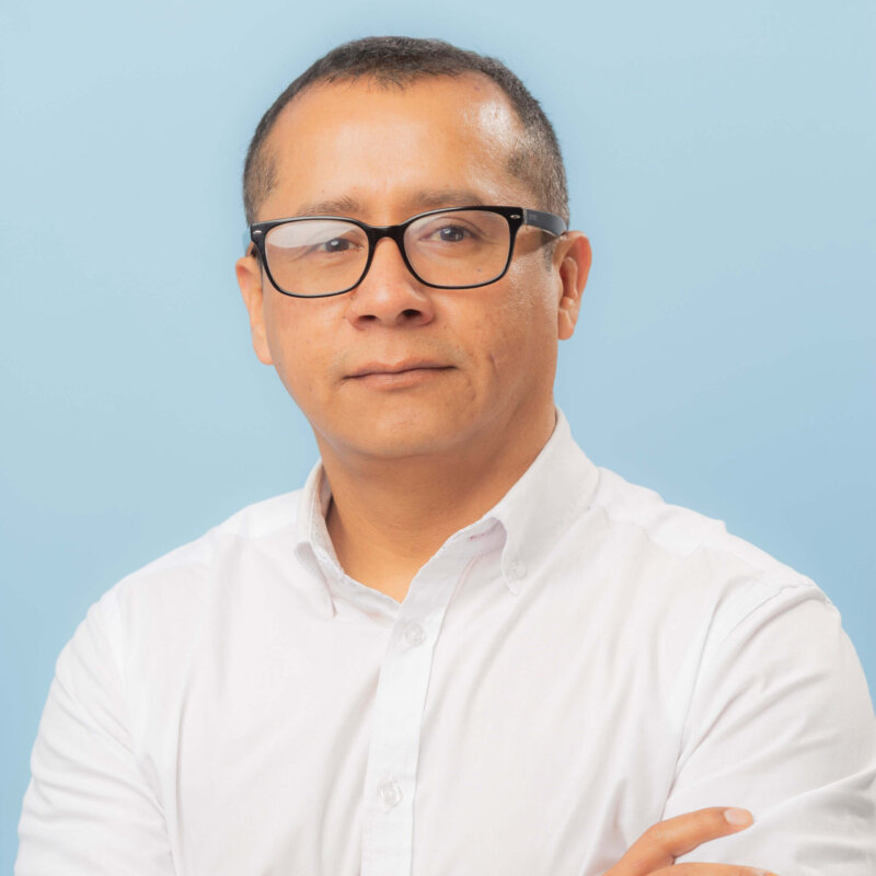 A man with short dark hair and glasses, wearing a white button-up shirt, stands with arms crossed against a light blue background.