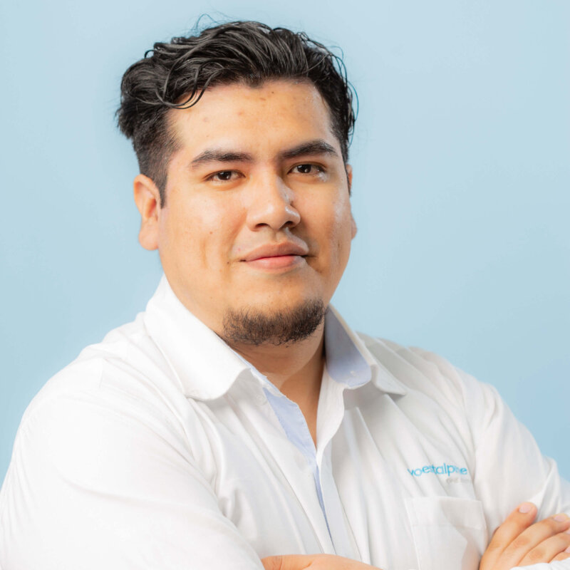 A man with short dark hair and a goatee, wearing a white collared shirt, stands against a light blue background with his arms crossed, looking confidently at the camera.