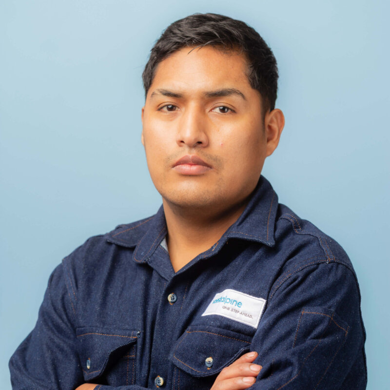 A man wearing a dark denim work shirt with a company patch stands against a light blue background, looking serious with his arms crossed, ready for contacto with clients.