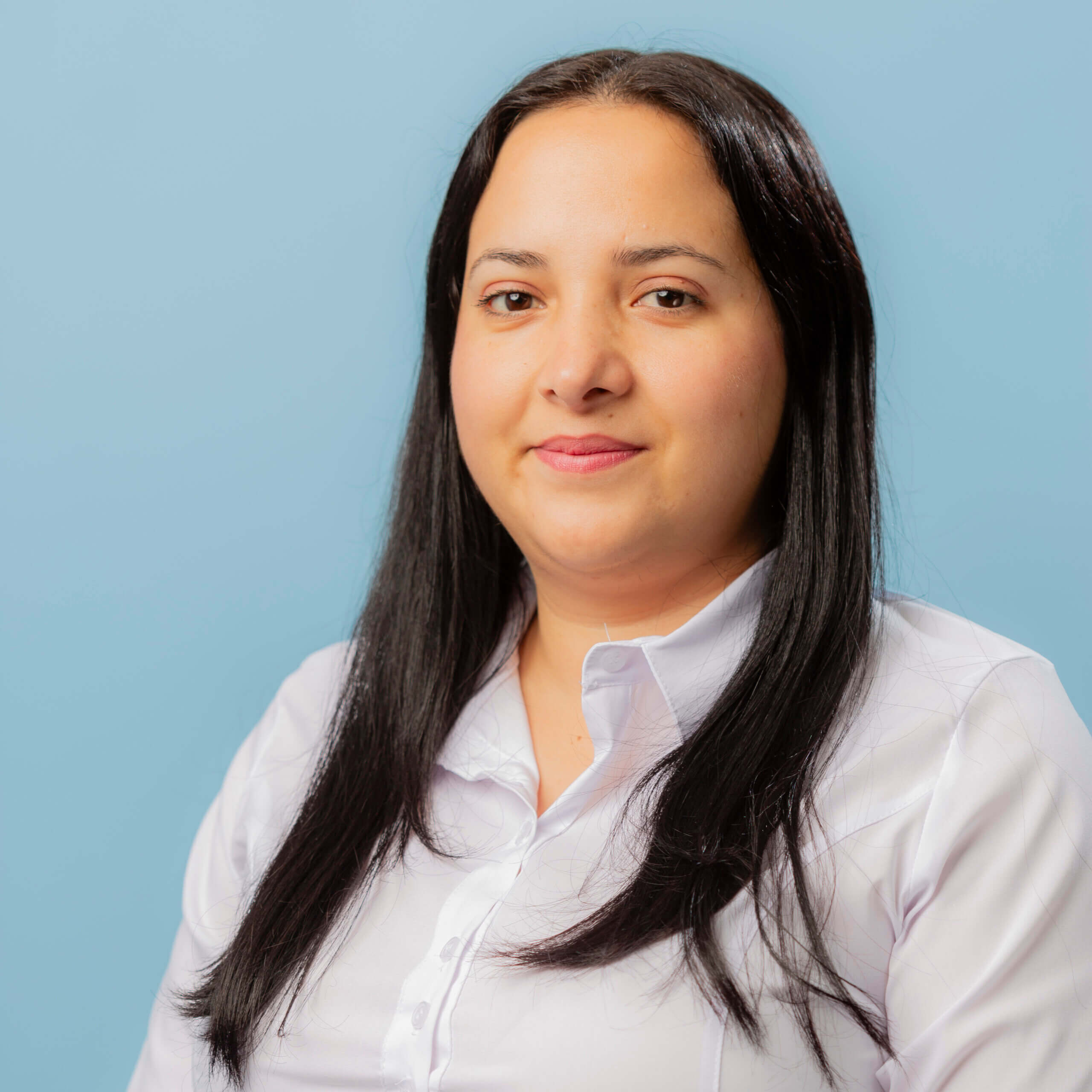 A woman with long straight black hair wearing a white blouse is posed in front of a plain light blue background, looking at the camera with a neutral expression, as if waiting for contacto.