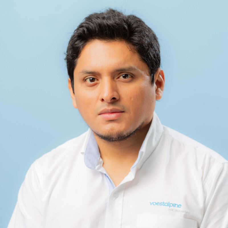 A man with short dark hair and light facial hair wears a white shirt with a blue voestalpine logo, posing against a plain light blue background, ideal for contacto or professional networking purposes.