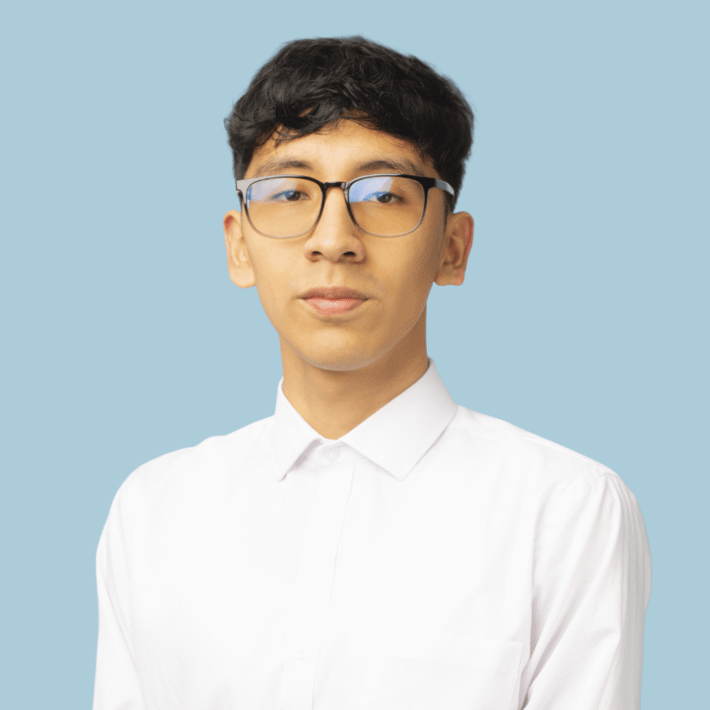 A young person with short dark hair and glasses, wearing a white button-up shirt, stands in front of a plain light blue background, ready to make contacto with those around them.