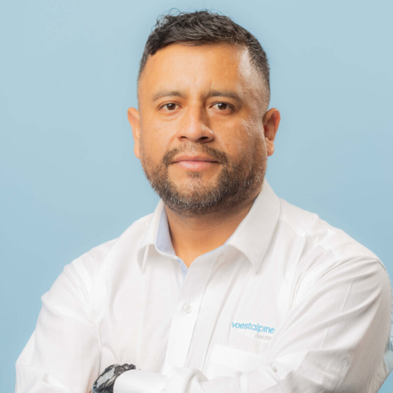 A man with short dark hair and a beard, wearing a white shirt with a voestalpine logo, stands against a light blue background with his arms crossed, looking at the camera with a neutral expression.