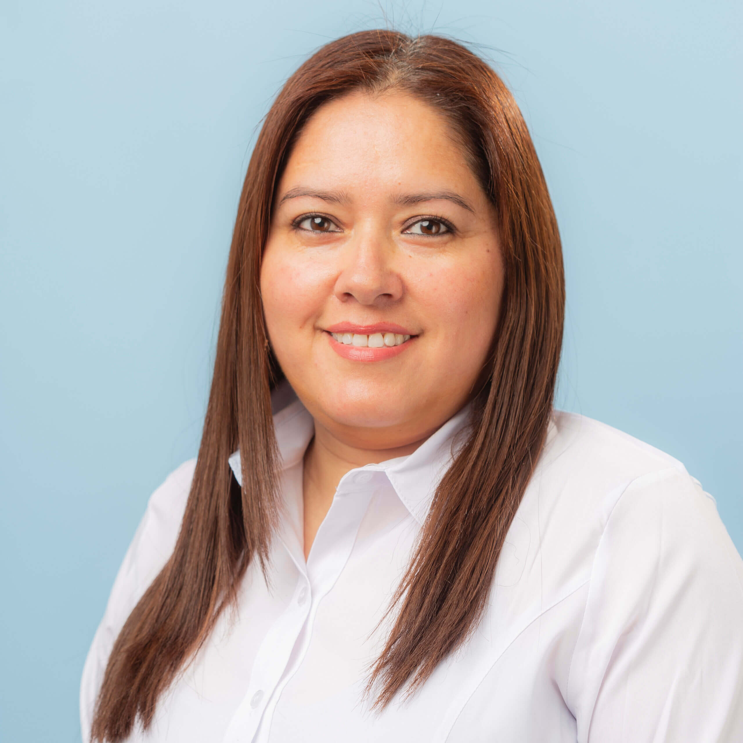 A woman with straight brown hair wearing a white shirt smiles at the camera against a light blue background, creating a warm sensación de contacto visual.
