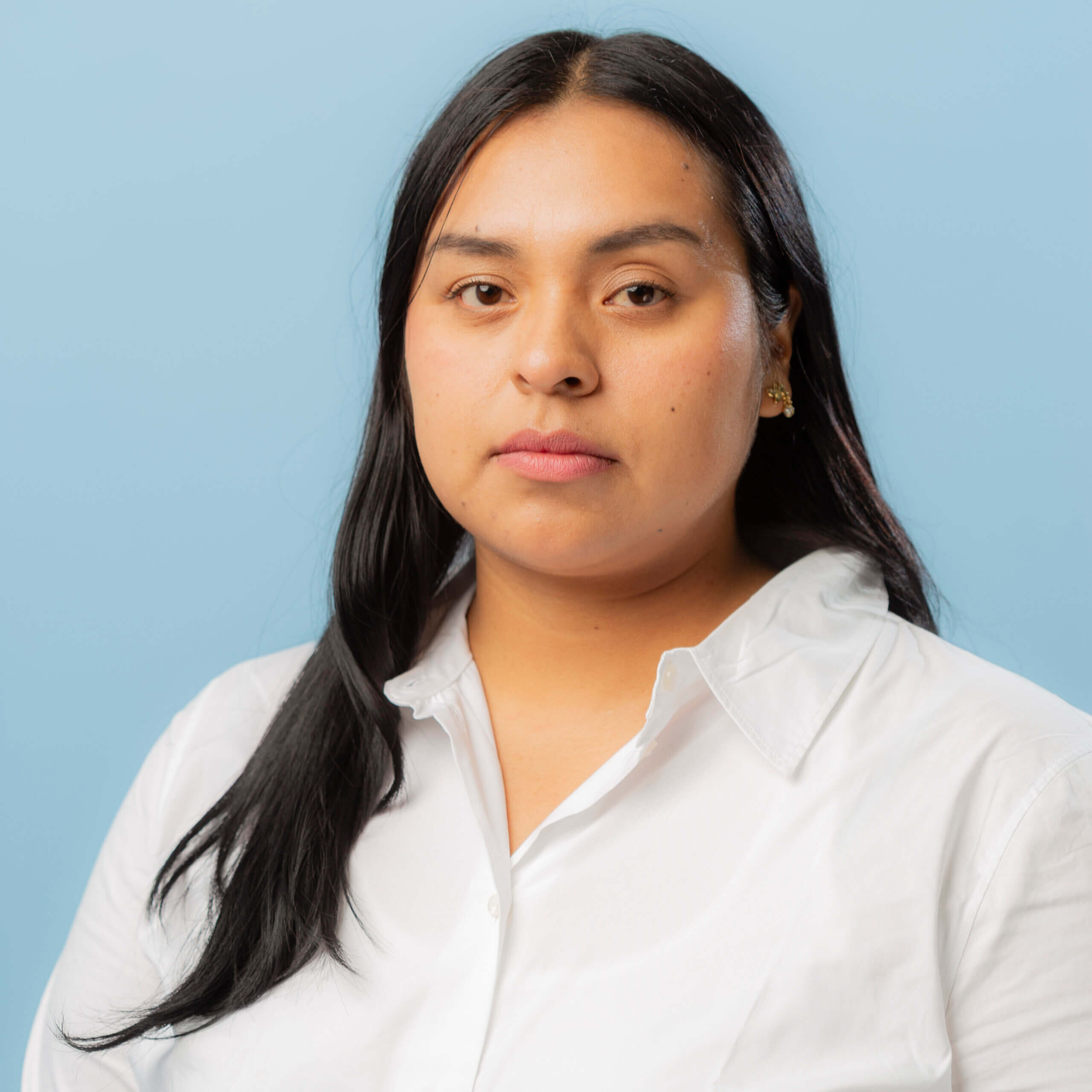 A woman with long black hair wearing a white collared shirt stands against a plain light blue background, looking directly at the camera with a neutral expression, as if ready for contacto or communication.