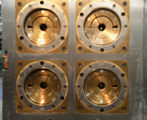 Close-up of a metallic industrial mold with four circular, gold-colored cavities, each with radial grooves and bolts around the edges, set in a square metal plate.