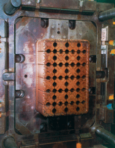 A close-up of a metal injection mold with multiple round holes arranged in a grid pattern on a rectangular copper insert, mounted inside a larger machine frame.
