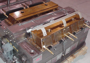 A close-up of a metal injection molding machine mold with several copper and steel components, hoses, and connectors, placed on a wooden pallet in an industrial setting.