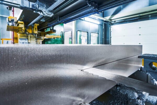 Close-up of an industrial metal cutting machine with a large, serrated saw blade slicing through a thick metal beam in a factory setting. Metal shavings are visible along the cut edge.