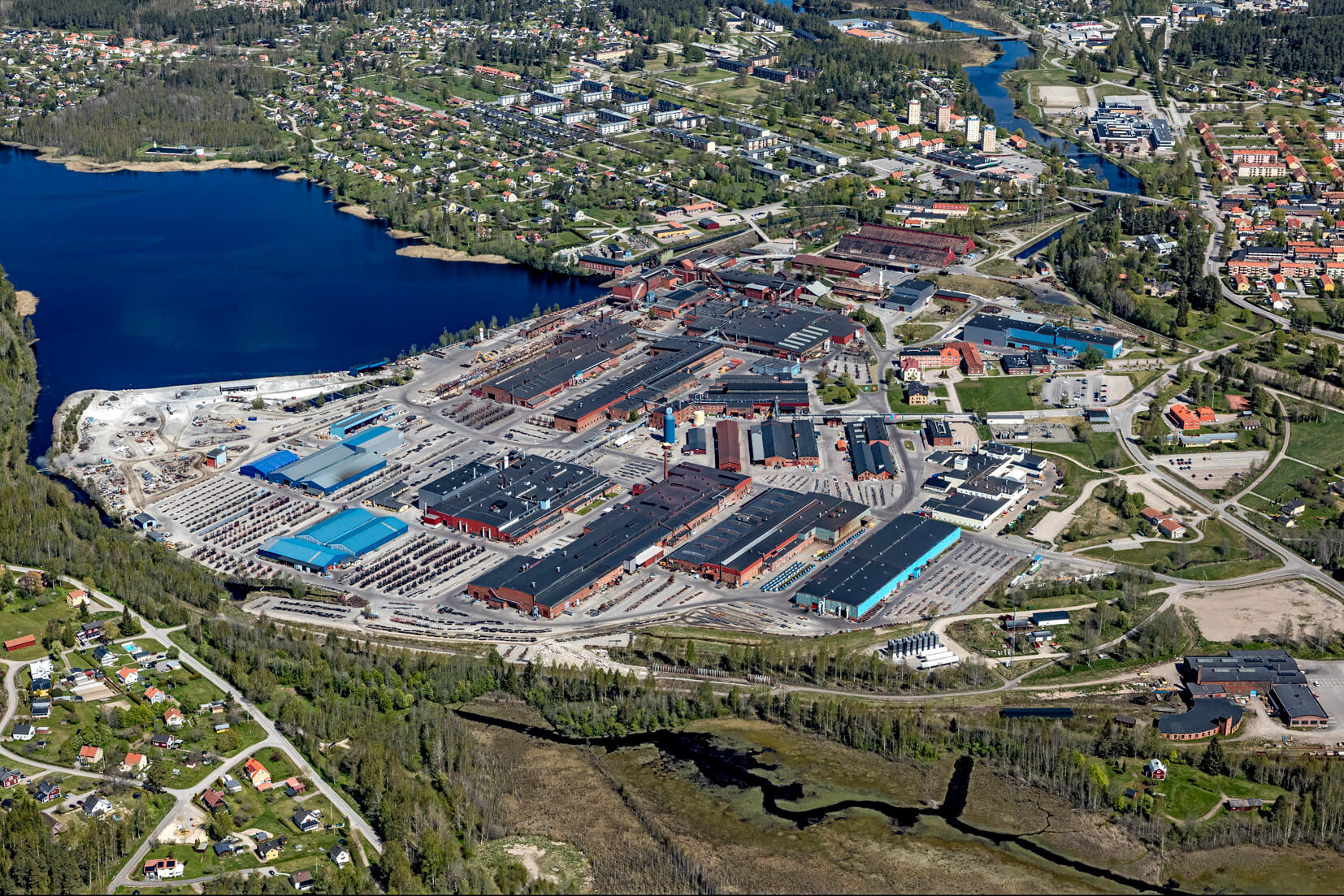 Aerial view of a large industrial complex with multiple buildings and blue-roofed sections, surrounded by trees, water, and a nearby residential area with houses and roads.